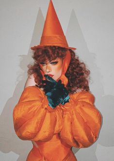 a woman in an orange dress and hat with her hands on her chest, posing for the camera