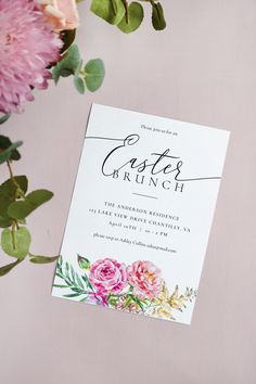 a wedding card with flowers and greenery on the front is laying next to a bouquet of pink carnations