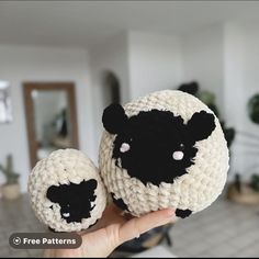 a hand holding two black and white crocheted sheeps in front of a living room