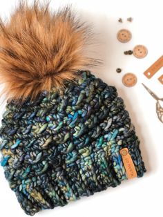 a knitted hat with a fur pom sits next to some buttons and scissors