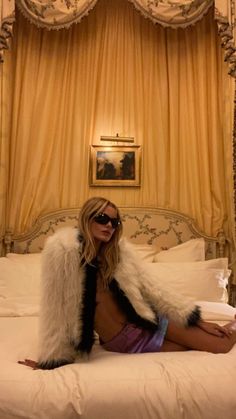 a woman sitting on top of a white bed in a room with yellow drapes