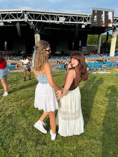 two women in white dresses standing on the grass at an outdoor music festival, holding hands