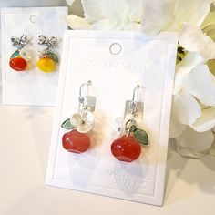 two pairs of earrings with fruit on them sitting next to a white flower and card