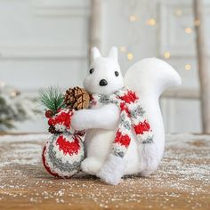 a white stuffed squirrel holding a pine cone in its paws and wearing a red and gray knitted scarf