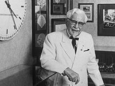 an old man in a white suit and bow tie is holding a cane near a wall with pictures on it