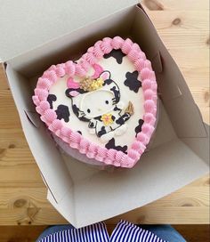 a hello kitty heart shaped cake in a box on a wooden table next to a person's hand