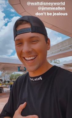 a man wearing a black shirt and hat smiles at the camera while holding his thumb up in front of him