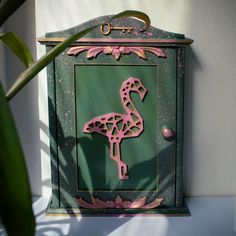 a pink flamingo painted on the side of a green mailbox next to a plant