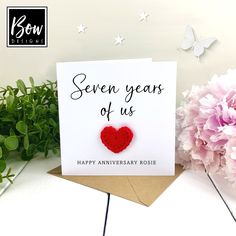 an anniversary card with a red heart on it next to some pink flowers and greenery