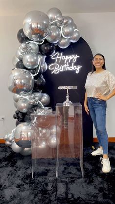 a woman standing in front of a happy birthday sign with balloons on the wall behind her