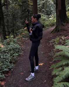 a woman standing on a path in the woods taking a photo with her cell phone