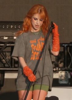 a woman with red hair and orange gloves on holding a microphone in front of a stage