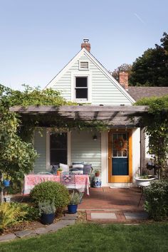 a white house with a table and chairs in the front yard, surrounded by greenery