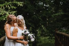 two brides embracing each other on a bridge in the woods with greenery behind them