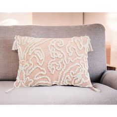 a light pink pillow on a grey couch with white trimmings and tasselled edges