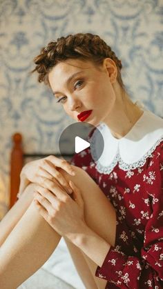 a woman sitting on top of a bed wearing a red dress and holding her hands together