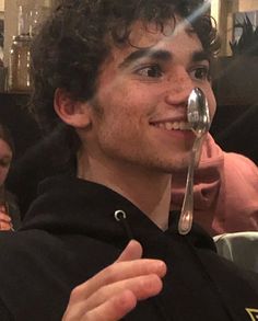 a young man with curly hair and spoon in his mouth smiles at the camera while sitting down