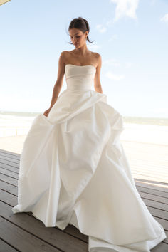 a woman in a white wedding dress is standing on a deck near the ocean and looking down