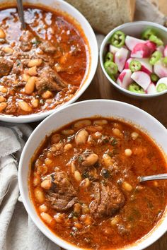 two bowls of soup with meat, beans and veggies next to a loaf of bread