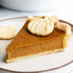 a slice of pumpkin pie on a plate with two cookies next to it and a bowl in the background