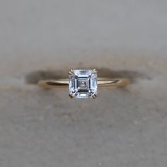 an emerald - cut diamond sits on top of a yellow gold engagement ring, which is sitting on concrete