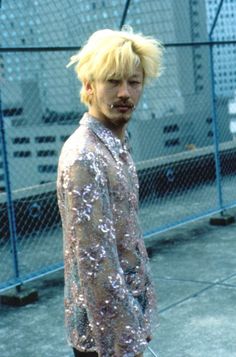 a man with blonde hair and piercings standing in front of a chain link fence