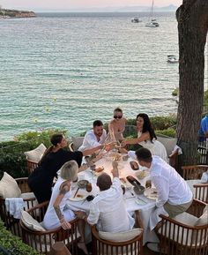 a group of people sitting around a table with food and drinks in front of the ocean