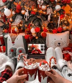 two people are holding cups with marshmallows in front of a christmas tree