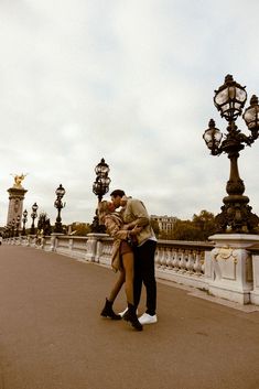 a man and woman kissing in front of some street lamps on a bridge over water