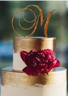 a gold and white wedding cake with a red flower on the bottom tier, topped with a monogram