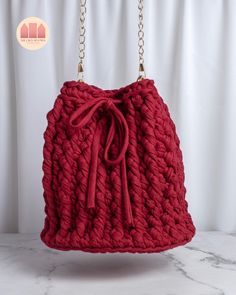 a red crocheted bag hanging from a chain on a marble counter top with white curtains in the background