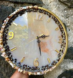 a hand holding a gold and white clock on top of a stone wall next to rocks