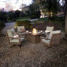 a fire pit surrounded by chairs and tables in the middle of a gravel area with trees