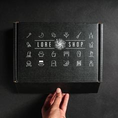 a person holding up a black box with white drawings on it that says lore shop