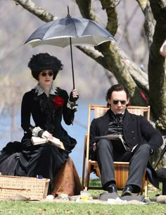 two people sitting on lawn chairs under an umbrella in the grass, with one person reading