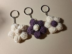 three crocheted flower keychains are shown on a white surface, one is purple and the other is white