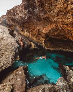 two people are swimming in the water near some large rocks and rock formations, while another person is diving into the pool