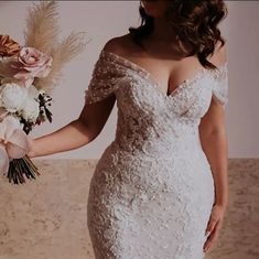 a woman in a white wedding dress holding a bouquet of flowers and wearing an off the shoulder gown