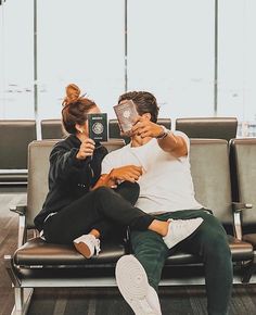 a man and woman sitting on a bench in an airport holding up their phones to take pictures