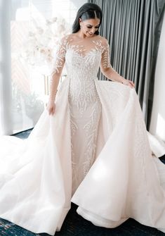 a woman in a wedding dress is standing near a window with her hand on the skirt