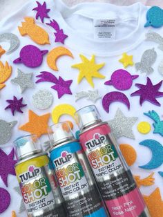 three cans of spray paint sitting on top of a white t - shirt with glitter stars and moon shapes