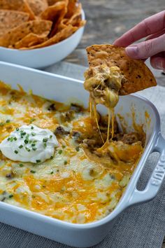 a person dipping a tortilla chip into a casserole dish with cheese and sour cream