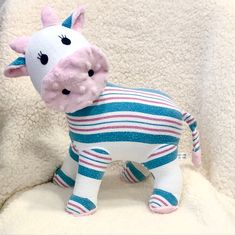 a stuffed cow is wearing a striped shirt and standing on a white blanket with its head turned to the side