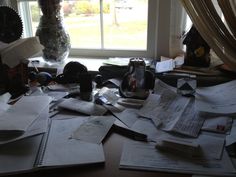 a messy desk with many papers on it and a vase full of flowers in the window