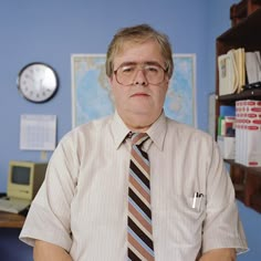 a man wearing glasses and a tie standing in front of a desk with a clock on the wall