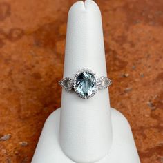 Estate/ Vintage 14KT White Gold Ring with Genuine, Oval Aquamarine + Diamond Accents and Halo Size 7.50, sizeable by us for a fee or your local jeweler Genuine, oval aquamarine measures: 9mm x7mm 1.51 CT Aquamarine Approx .20 CT natural diamonds; SI1/2 clarity Diamonds on the scrol and halo 14KT Gold Weighs 2.90 Grams Gold is patina'd and we like the look and leaving like this. Available to rhodium if desired An elegant piece with the square faceted Aquamarine center + sparkling, side diamonds! Oval Aquamarine Jewelry With Accent Stones, Oval Aquamarine Anniversary Jewelry, Oval Aquamarine Fine Jewelry, Oval Aquamarine Center Stone Jewelry, Oval Aquamarine Jewelry With Center Stone, Gia Certified Oval Blue Topaz Ring, Oval Aquamarine Light Blue Jewelry, Light Blue Oval Rings For Anniversary, Oval Aquamarine Ring With Diamond Accents