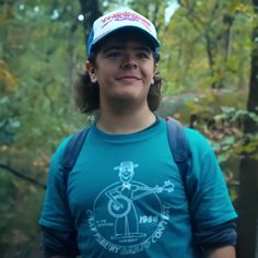 a woman in a blue shirt and hat standing in the woods with trees behind her