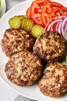 a white plate topped with hamburger patties next to veggies and pickles