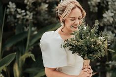 a woman in a white dress holding a bouquet of greenery and smiling at the camera