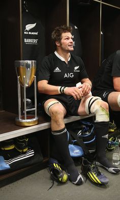 two rugby players are sitting in the locker room with their socks on and one is holding a trophy